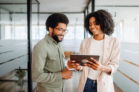 Two cheerful colleagues are sharing a tablet screen, the woman pointing at the screen, a man watching with interest, collaborative and friendly work atmosphere. Team interaction and knowledge exchange