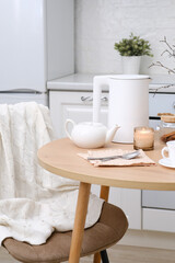Fototapeta na wymiar Home spring interior in kitchen. Beige chair with white cover, white electric kettle, hot teapot, candle, cup of tea on round wooden table in cosy white kitchen.