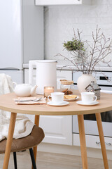 Home spring easter interior in kitchen. Beige chair with white cover, white electric kettle, hot teapot, vase with spring branches candle, cups of tea on round wooden table in cosy white kitchen.