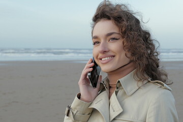 Beautiful young smiling woman talking on the cell phone on the beach with ocean and sky in the...