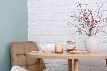 Home spring easter design in kitchen. White teapot, plate with cookies, ceramic vase with spring branches on round wooden table, beige chair with cover on green background in cosy living room..