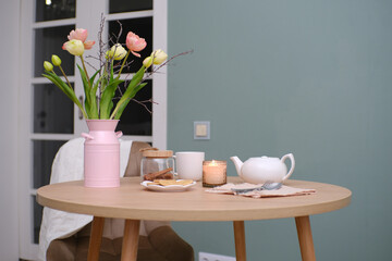 Home spring interior in living room. Pink vase with tulips and branches,hot teapot, burning candle, cup of tea on round wooden table in cosy living room.