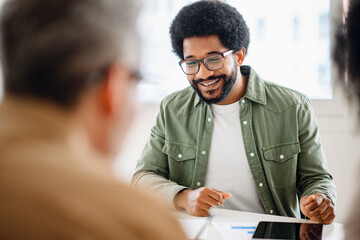 African-American man with a warm smile, wearing glasses and a green casual shirt, is engaged in conversation with an out-of-focus colleague, suggesting an approachable and friendly office discussion - obrazy, fototapety, plakaty