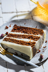 A piece of carrot cake served on rustic plate placed on bright sunny scene. Layered cake with cream cheese frosting and fluffy biscuit. Served with tea. 