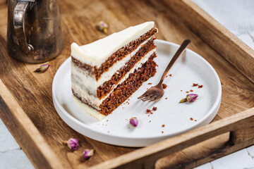 A piece of carrot cake served on rustic plate placed on bright sunny scene. Layered cake with cream cheese frosting and fluffy biscuit. Served with tea. 