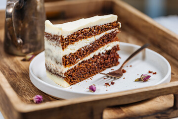 A piece of carrot cake served on rustic plate placed on bright sunny scene. Layered cake with cream cheese frosting and fluffy biscuit. Served with tea. 