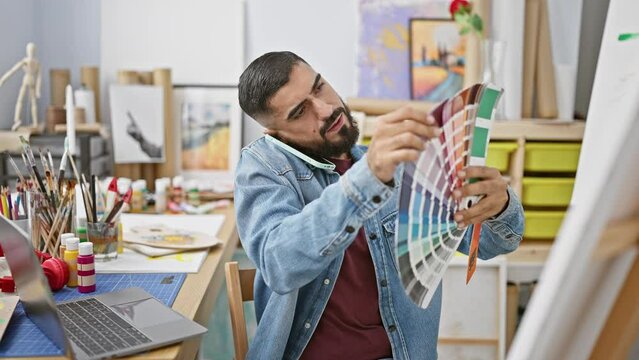 Bearded man examines color swatches in a creative studio with art supplies and a laptop, embodying design and creativity.
