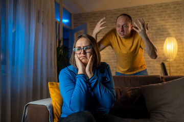 Portrait of tired stressed woman sadness looking away sitting on bed on blurred background of aggressive husband shouting on wife at home. Concept of family scandal, crisis, domestic violence, abuse. - obrazy, fototapety, plakaty