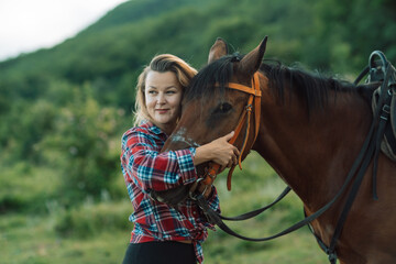 Obraz premium Happy blonde with horse in forest. Woman and a horse walking through the field during the day. Dressed in a plaid shirt and black leggings.