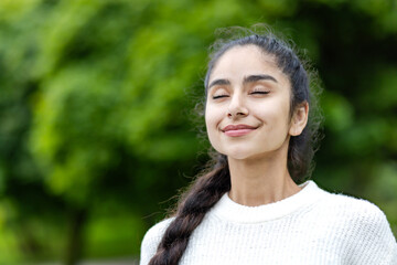 A young beautiful Indian woman is standing outside in a park in a white sweater and with her eyes...