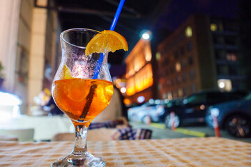 Aperolspritz with lemon and a straw against the backdrop of the evening city.