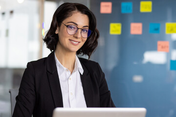 Calm busy woman in eyeglasses looking at camera with smile while sitting in workspace with coloruful stickers on wall. Pretty hospitable secretary in shirt meeting business partners of company ceo.