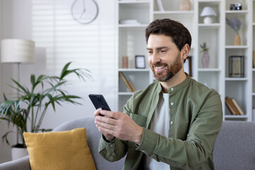 Happy bearded man enjoying using cellular phone while sitting on couch in living room. Careless...