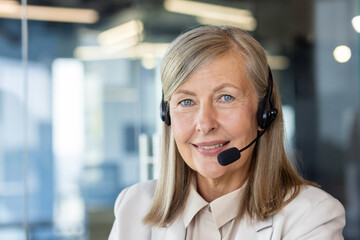 Portrait of blonde mature female in wireless headset with microphone smiling on blurred background. Successful head of consulting company getting ready for online communication with business client.