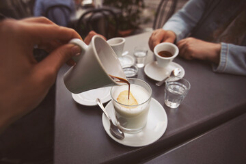 Woman hand pouring coffee into a glass of milk. friends hands holding cups of coffee on wooden...