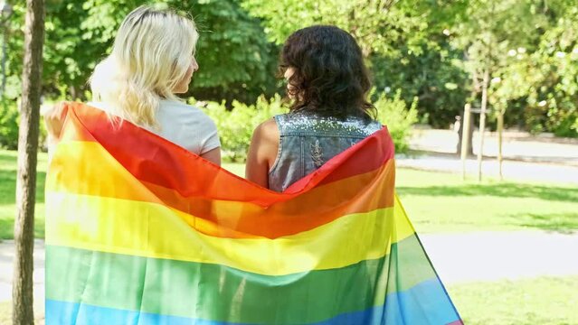 lesbian couple looking at each other walking through a park with their backs to each other with a gay pride flag on their shoulders