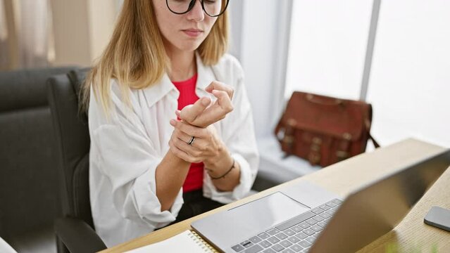 A young woman experiencing wrist pain at her office desk with a laptop and smartphone visible.