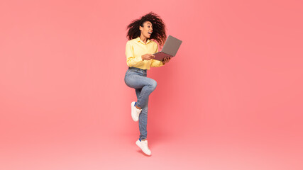 Excited black woman jumps, holding laptop on pink background