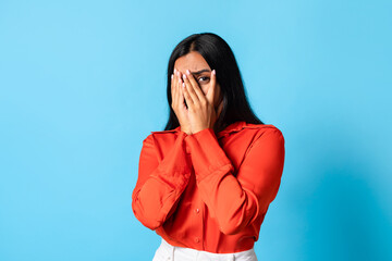 scared young indian woman peeking through fingers against blue backdrop