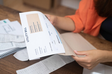 stern woman meticulously reviews utility bills, pondering rising costs of electricity, water, and...