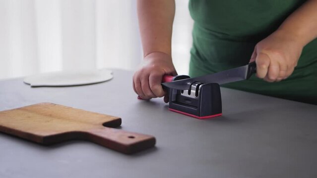 A housewife woman sharpens a kitchen knife in a knife sharpener on the table. Slowmotion