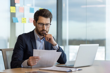 Serious thinking businessman behind paper work, mature experienced boss working with reports and financial documents, financier inside office with laptop, reading contract, thoughtfully checking.