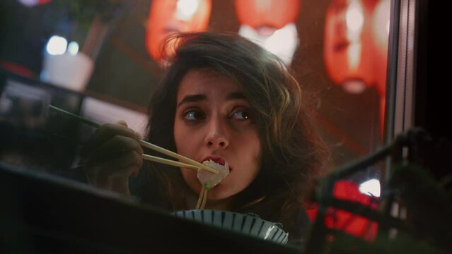 Reflective woman enjoys Chinese meal with chopsticks observing night city street through window. Appealing lady takes well-deserved break after workday in Asian restaurant during late evening