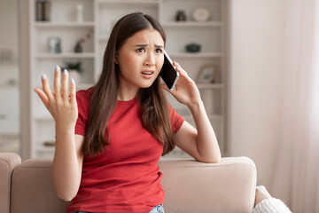 Perplexed young asian woman gesturing with her hand while talking on phone - obrazy, fototapety, plakaty