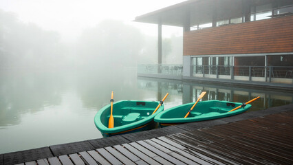A lake with a cafe building near the water in the fog. In the foreground, a pier with paddle boats wet from rain. Trees are reflected in the water. Copy space.