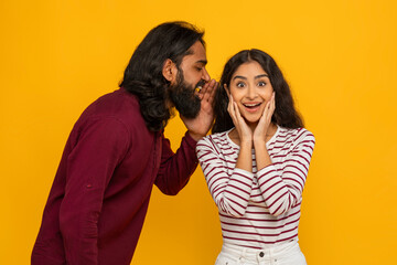 Young indian guy sharing secret with his girlfriend, yellow background
