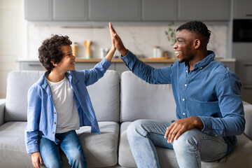Joyful black father and son high-fiving, celebrating happy moment on the couch