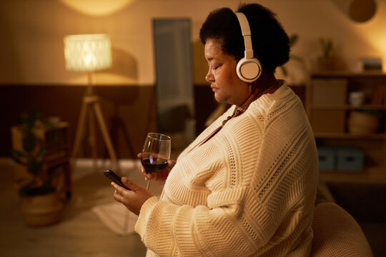 Side View Portrait Of Senior Black Woman Using Smartphone While Drinking Wine At Home And Listening To Music With Headphones