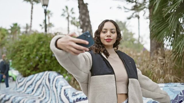 Young woman takes a selfie in a park with palm trees, showcasing urban lifestyle and technology use outdoors.