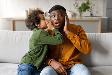 Son whispers secret to surprised black father on the couch - obrazy, fototapety, plakaty