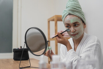 Skincare Retreat: A bathrobe-clad beauty uses a brush to apply a clay mask, admiring her reflection during her organic facial treatment. 