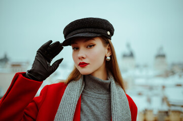 Fashionable woman wearing cap, red winter coat, gray scarf, turtleneck sweater, leather gloves, posing on balcony with beautiful view on snow covered European city. Copy, empty, blank space for text