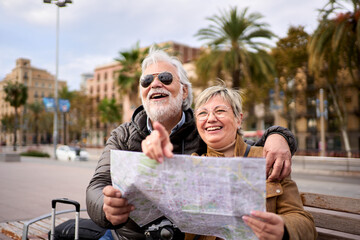 Senior Caucasian tourist couple smiling with travel map in hands looking and pointing interest places. Elderly husband and wife sitting on bench in city street enjoying vacation. Adult tourism people 