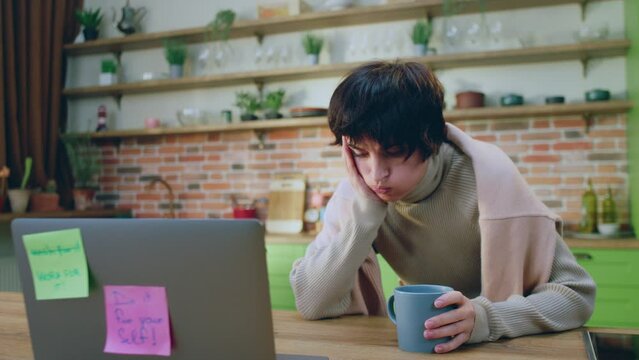 Very tired and bad feeling woman in the morning holding her cup of tea at kitchen island she looking over her laptop at home