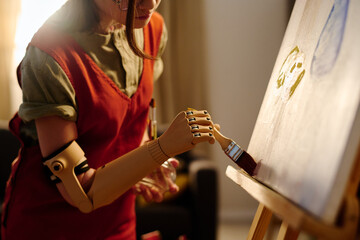 Myoelectric hand of amputee schoolgirl with paintbrush working over new painting while standing in...