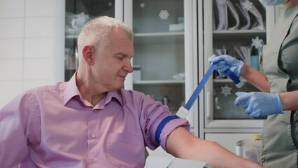 Nurse in professional uniform places tourniquet to identify viable and accessible vein on senior man arm. Experienced doctor in rubber gloves ties tourniquet preparing for blood sample collection