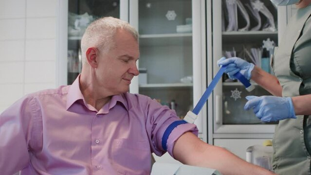 Nurse in professional uniform places tourniquet to identify viable and accessible vein on senior man arm. Experienced doctor in rubber gloves ties tourniquet preparing for blood sample collection