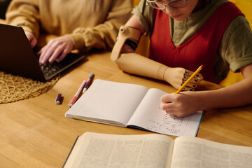 Cropped shot of diligent schoolgirl with myoelectric arm making notes in copybook while sitting by...