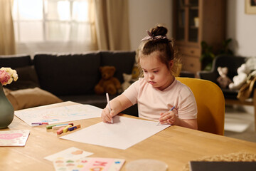 Cute little girl with mental disability sitting by table in front of camera and drawing with...