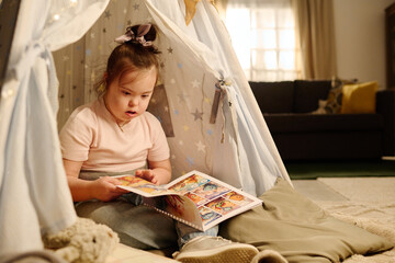 Sweet girl with Down syndrome sitting on the floor in tent and looking at pictures in book of...