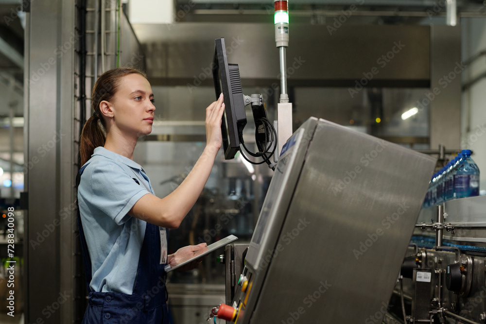Aufkleber Young female inspector with tablet standing in front of factory equipment and entering data while touching interactive display