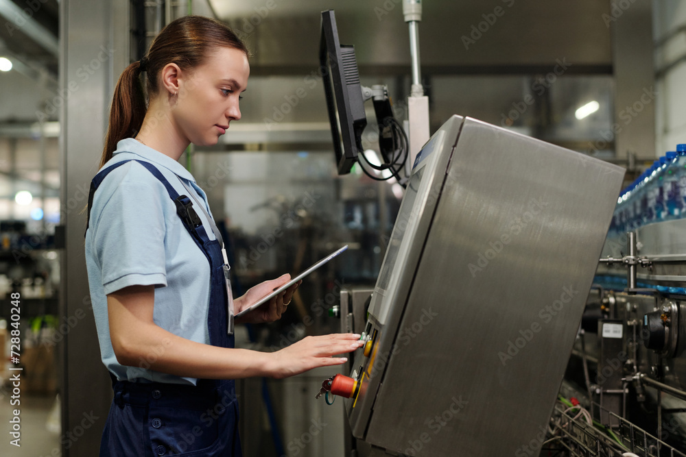 Poster young worker of plant standing in front of factory equipment and pressing button on control panel of