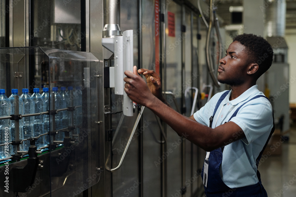 Poster young african american worker touching display on panel of automated equipment with row of capped pl