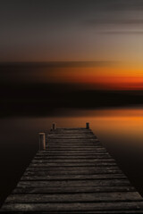 Fototapeta na wymiar Abstract lake landscape at sunset with an old wooden boardwalk. Dock in lake with abstract blurred background in dark sunset evening