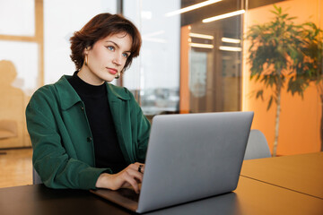 Focused DevOps Engineer, a woman, reads data on her computer screen in a modern office,...