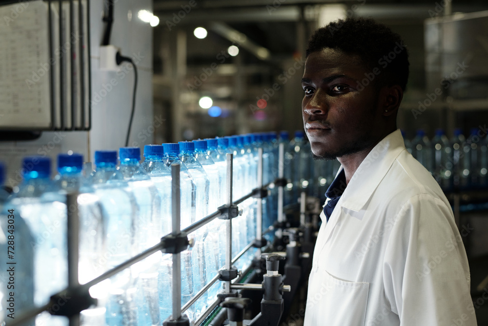 Poster Young African American quality control engineer in labcoat standing in front of assembly line with capped bottled drinks
